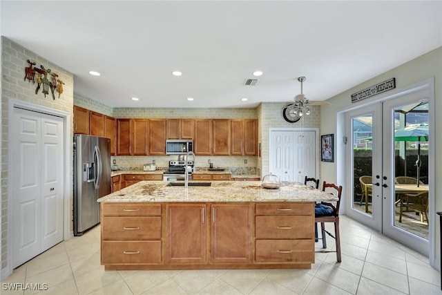 kitchen with ceiling fan, a kitchen island with sink, appliances with stainless steel finishes, french doors, and light stone counters