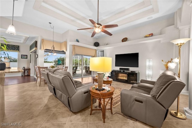 living area with light tile patterned floors, a ceiling fan, a glass covered fireplace, ornamental molding, and a tray ceiling