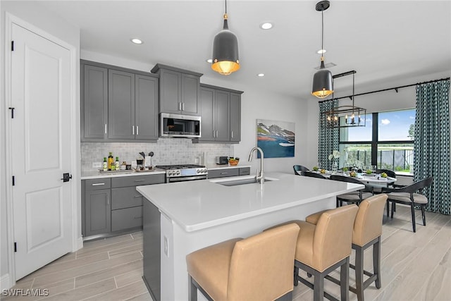 kitchen with stainless steel appliances, sink, a center island with sink, gray cabinets, and hanging light fixtures