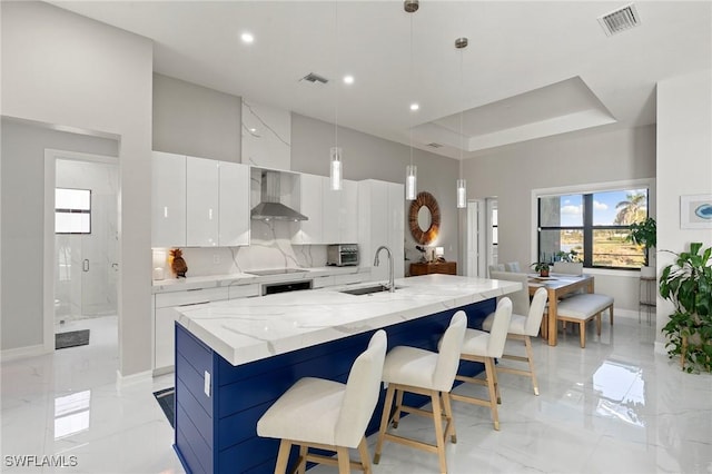 kitchen featuring sink, wall chimney range hood, decorative light fixtures, a center island with sink, and white cabinets