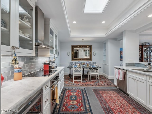 kitchen featuring a raised ceiling, wall chimney range hood, hanging light fixtures, sink, and appliances with stainless steel finishes