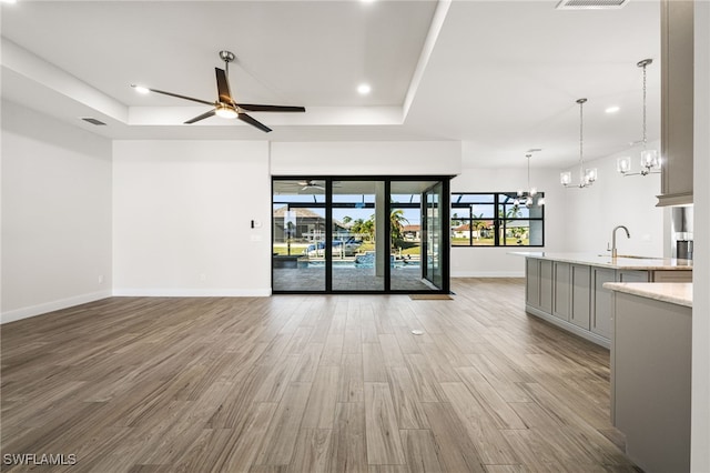 unfurnished living room with a tray ceiling, sink, ceiling fan with notable chandelier, and light hardwood / wood-style flooring