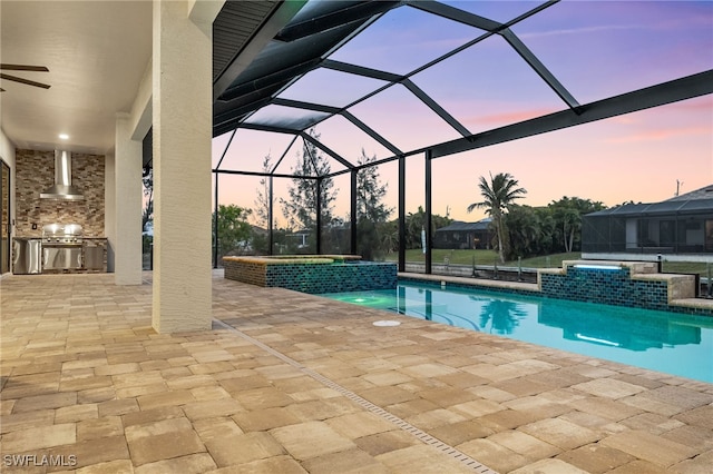 pool at dusk with an in ground hot tub, pool water feature, glass enclosure, an outdoor kitchen, and a patio