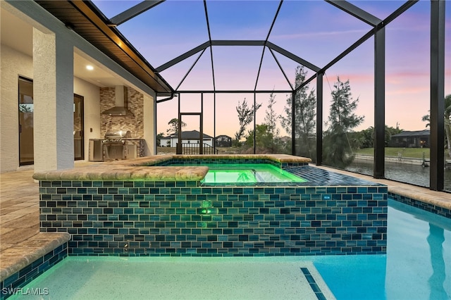 pool at dusk with glass enclosure, exterior kitchen, a water view, an in ground hot tub, and a patio