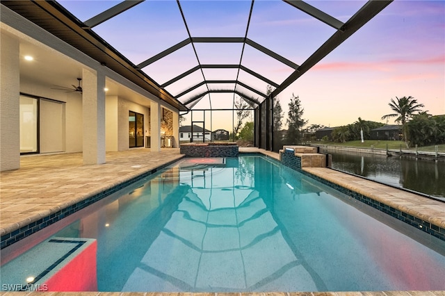 pool at dusk featuring a water view, ceiling fan, glass enclosure, and a patio area