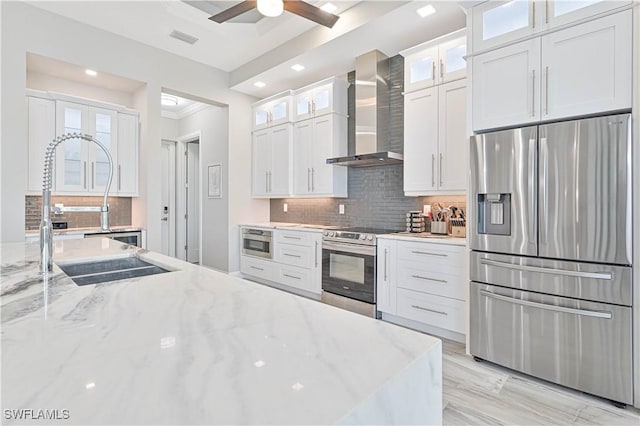 kitchen with wall chimney range hood, white cabinetry, backsplash, stainless steel appliances, and light stone counters