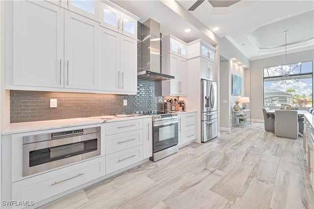 kitchen featuring a raised ceiling, white cabinetry, tasteful backsplash, stainless steel appliances, and wall chimney exhaust hood