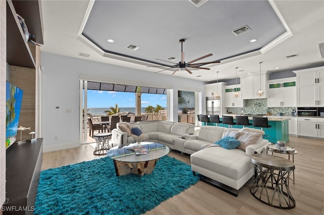 living room with a raised ceiling, ceiling fan, and light wood-type flooring