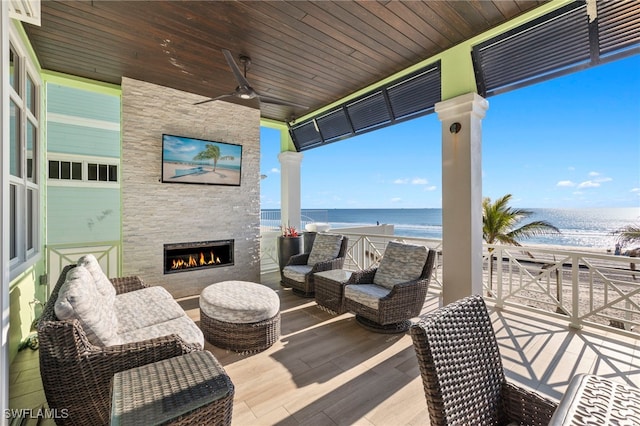 wooden terrace with a view of the beach, an outdoor living space with a fireplace, ceiling fan, and a water view