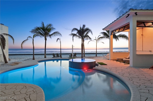 pool at dusk with a patio area, an in ground hot tub, and a water view