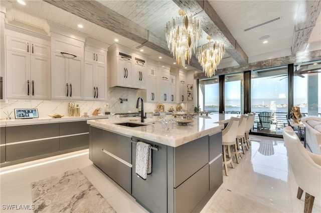 kitchen featuring a notable chandelier, a center island with sink, beamed ceiling, white cabinetry, and light stone countertops