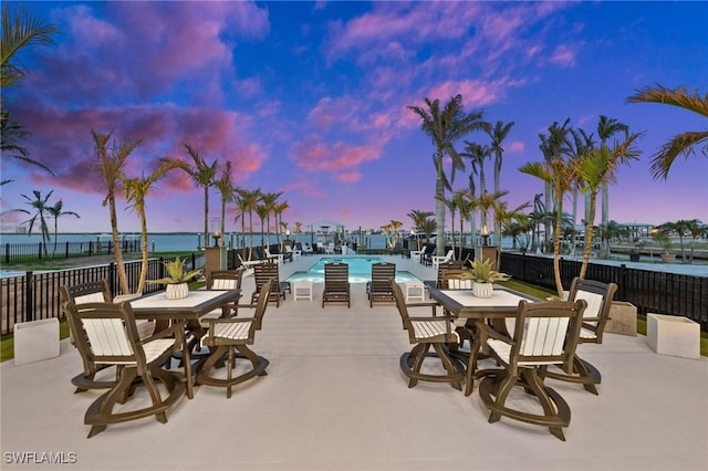 patio terrace at dusk featuring a community pool, fence, and a water view
