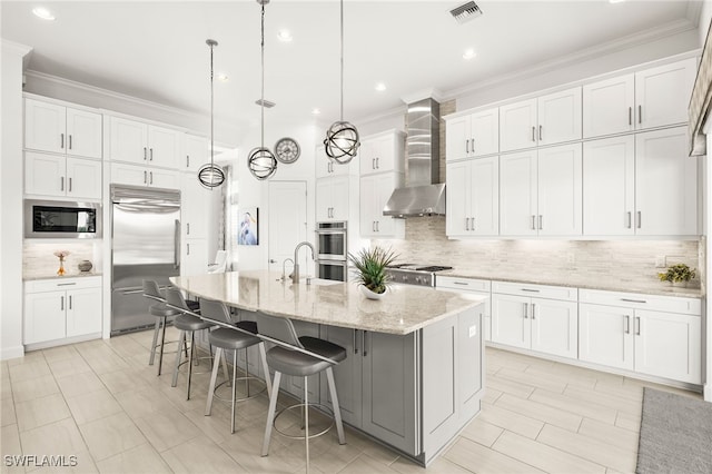 kitchen with built in appliances, white cabinetry, a center island with sink, and wall chimney range hood