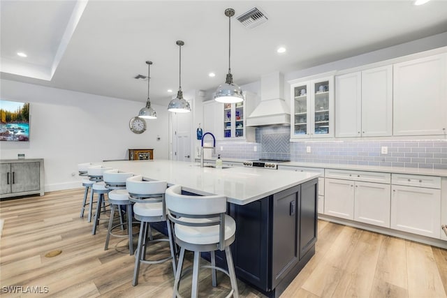 kitchen featuring white cabinetry, sink, premium range hood, pendant lighting, and a center island with sink