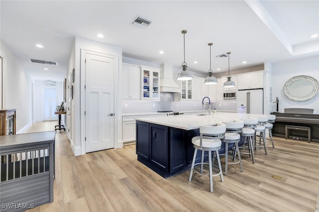 kitchen featuring custom range hood, a kitchen island with sink, white fridge with ice dispenser, white cabinetry, and hanging light fixtures