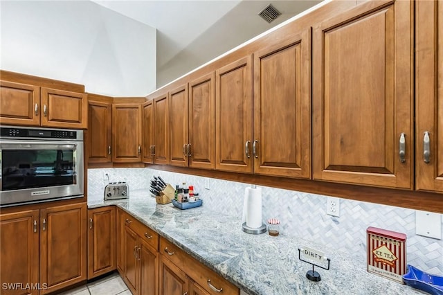 kitchen with decorative backsplash, stainless steel oven, light stone counters, and brown cabinets