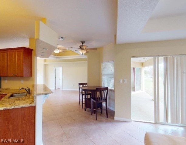 kitchen with ceiling fan, sink, light stone counters, kitchen peninsula, and a tray ceiling