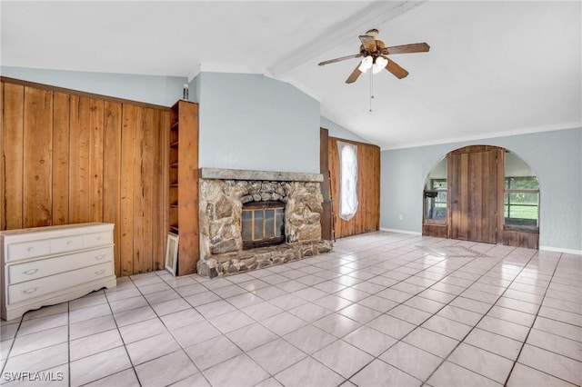 unfurnished living room with ceiling fan, wooden walls, light tile patterned floors, lofted ceiling with beams, and a stone fireplace