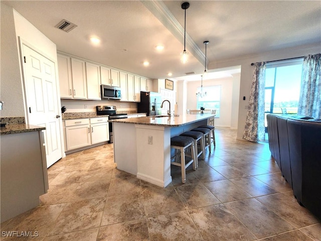 kitchen with white cabinets, appliances with stainless steel finishes, decorative light fixtures, stone countertops, and a center island with sink