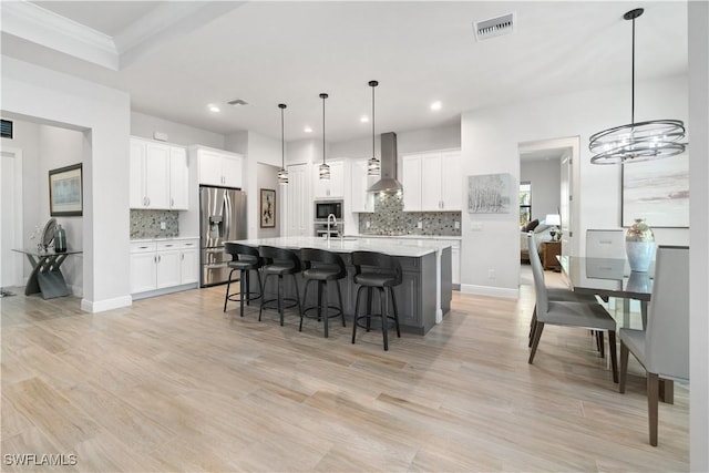 kitchen featuring white cabinetry, wall chimney range hood, an island with sink, hanging light fixtures, and stainless steel fridge with ice dispenser