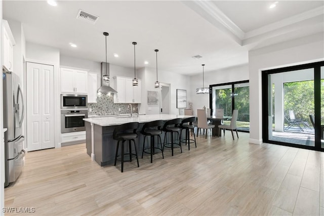 kitchen with pendant lighting, a kitchen island with sink, appliances with stainless steel finishes, white cabinets, and wall chimney exhaust hood