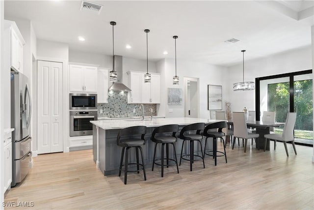 kitchen with pendant lighting, wall chimney range hood, appliances with stainless steel finishes, and a center island with sink