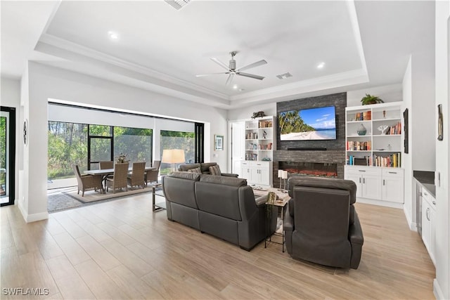 living room featuring light hardwood / wood-style floors, a stone fireplace, ceiling fan, a tray ceiling, and built in shelves