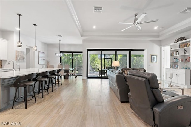 living room with ceiling fan, sink, a tray ceiling, crown molding, and light wood-type flooring