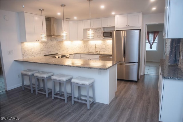 kitchen with appliances with stainless steel finishes, wall chimney exhaust hood, white cabinetry, hanging light fixtures, and kitchen peninsula