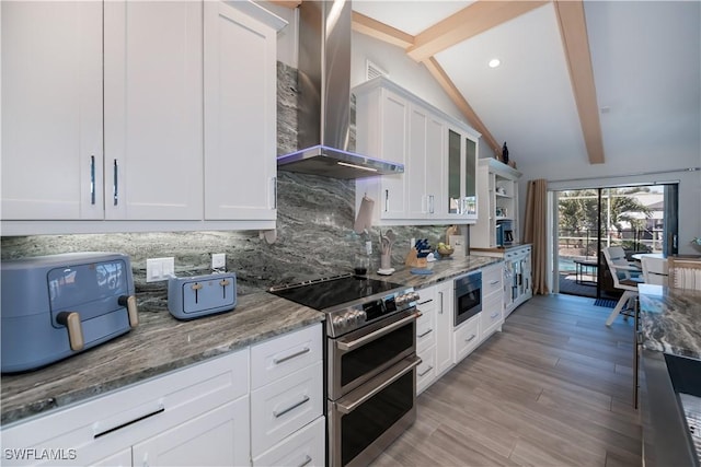 kitchen featuring wall chimney exhaust hood, vaulted ceiling with beams, white cabinetry, stainless steel appliances, and decorative backsplash