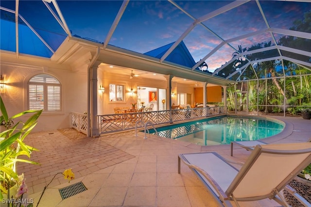 pool at dusk with glass enclosure, a patio area, and ceiling fan