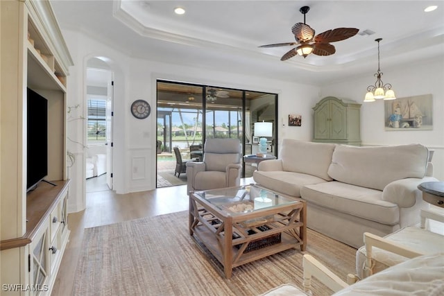 living room with ceiling fan with notable chandelier, a raised ceiling, light wood-type flooring, and crown molding