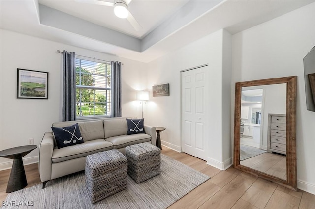 living room featuring ceiling fan, light wood finished floors, a raised ceiling, and baseboards