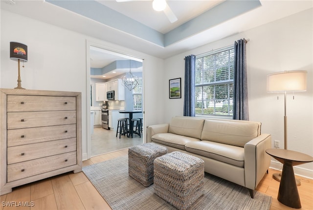 living room with ceiling fan with notable chandelier, light wood-type flooring, a raised ceiling, and baseboards