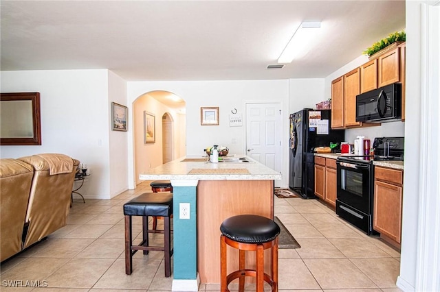 kitchen featuring light tile patterned flooring, a kitchen bar, an island with sink, and black appliances