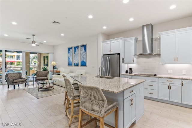 kitchen with wall chimney exhaust hood, a kitchen island with sink, ceiling fan, white cabinetry, and stainless steel refrigerator