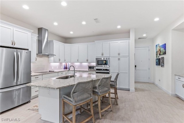 kitchen with sink, wall chimney exhaust hood, a kitchen island with sink, white cabinets, and appliances with stainless steel finishes