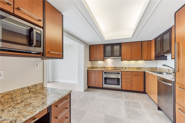 kitchen with light stone counters, a tray ceiling, appliances with stainless steel finishes, glass insert cabinets, and a sink