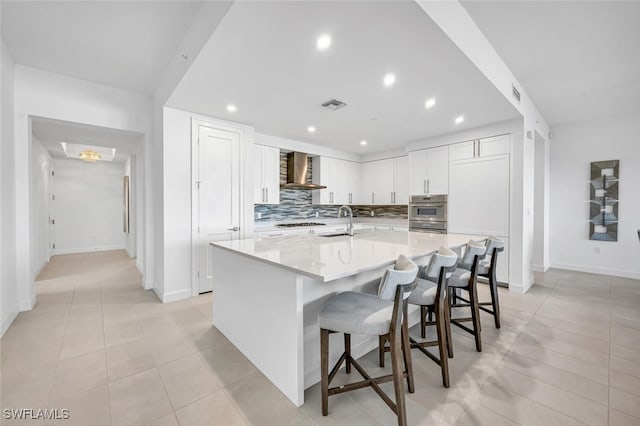 kitchen featuring wall chimney exhaust hood, a breakfast bar area, tasteful backsplash, an island with sink, and white cabinets