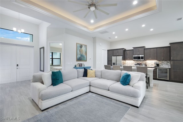 living room featuring light wood-type flooring, ceiling fan with notable chandelier, a raised ceiling, and beverage cooler