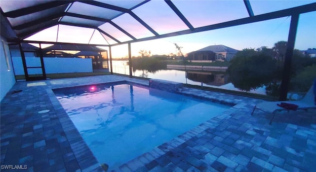 pool at dusk featuring glass enclosure, a water view, and a patio area