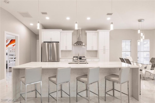 kitchen with wall chimney exhaust hood, hanging light fixtures, stainless steel appliances, white cabinetry, and a large island with sink