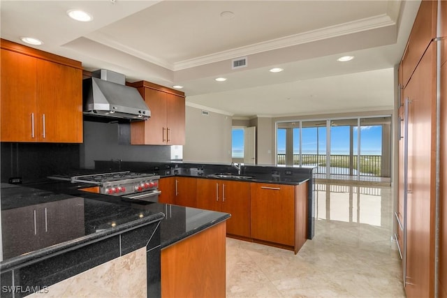 kitchen with kitchen peninsula, sink, dark stone counters, a tray ceiling, and wall chimney exhaust hood