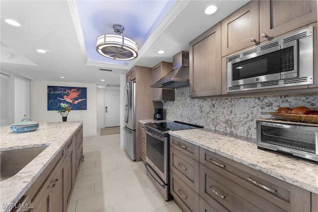 kitchen featuring appliances with stainless steel finishes, wall chimney exhaust hood, decorative backsplash, a raised ceiling, and light stone counters