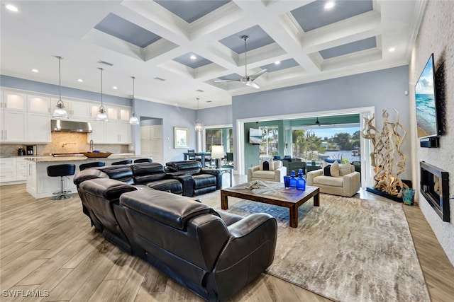 living room with ceiling fan, beamed ceiling, coffered ceiling, light wood-type flooring, and a stone fireplace