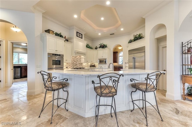 kitchen featuring a spacious island, decorative backsplash, built in appliances, a breakfast bar area, and light stone counters