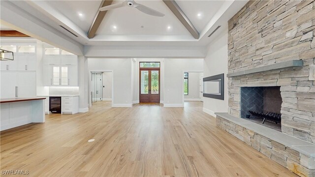 unfurnished living room with wine cooler, ceiling fan, a towering ceiling, a stone fireplace, and light hardwood / wood-style floors