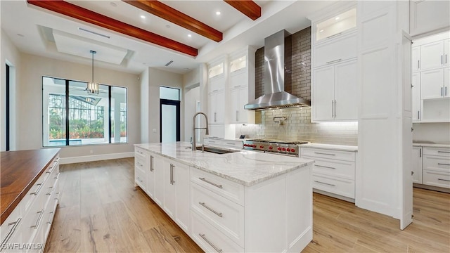 kitchen with stainless steel stove, decorative backsplash, wall chimney range hood, white cabinets, and an island with sink