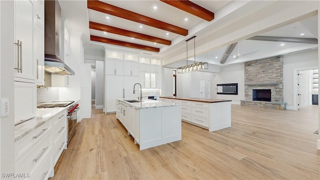 kitchen with hanging light fixtures, white cabinets, tasteful backsplash, a kitchen island with sink, and wall chimney range hood