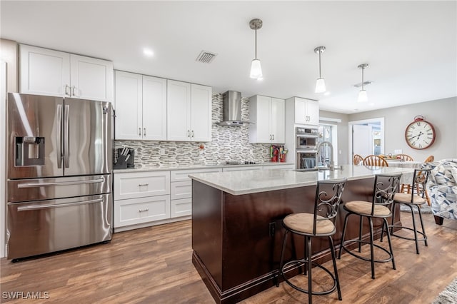 kitchen with white cabinetry, stainless steel appliances, a kitchen island with sink, hanging light fixtures, and wall chimney exhaust hood